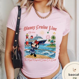 K&ouml;nnte beinhalten: Hellrosa T-Shirt mit einem Aufdruck von Minnie Mouse auf einem Kreuzfahrtschiff. Das Design beinhaltet den Text "Disney Cruise Line Est. 1998" und "The Most Magical Cruise On Earth", einen Leuchtturm, Palmen und Feuerwerk.