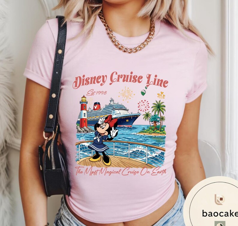 K&ouml;nnte beinhalten: Hellrosa T-Shirt mit einem Aufdruck von Minnie Mouse auf einem Kreuzfahrtschiff. Das Design beinhaltet den Text "Disney Cruise Line Est. 1998" und "The Most Magical Cruise On Earth", einen Leuchtturm, Palmen und Feuerwerk.