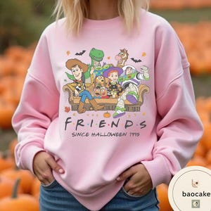May include: A light pink long-sleeve sweatshirt featuring a graphic print of Toy Story characters sitting on a couch with the text "FRIENDS SINCE HALLOWEEN 1995". The design includes Woody, Buzz Lightyear, Jessie, Rex, Bullseye, and Slinky Dog.