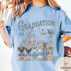 May include: Light blue t-shirt with the word "Graduation" in a vintage font. The graphic features Disney characters in graduation attire, standing in front of a castle and other buildings. The design includes graduation caps, diplomas, and books.