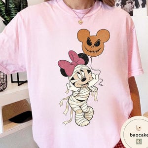 May include: A light pink t-shirt featuring a cartoon mummy character with a pink bow and a jack-o'-lantern balloon. The mummy is wrapped in bandages and has a smiling expression. The balloon is brown with a carved face.