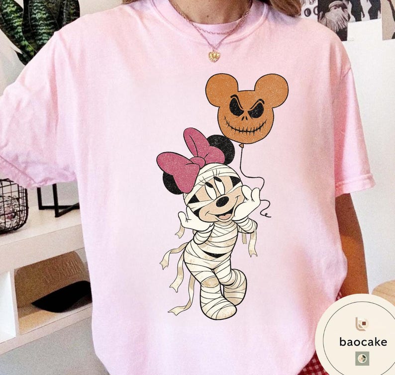 May include: A light pink t-shirt featuring a cartoon mummy character with a pink bow and a jack-o'-lantern balloon. The mummy is wrapped in bandages and has a smiling expression. The balloon is brown with a carved face.