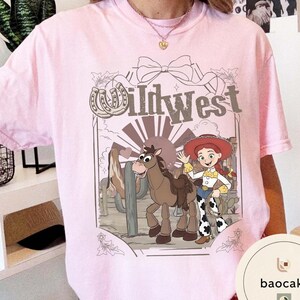 Pu&ograve; includere: T-shirt rosa chiaro con un design a cartoni animati a tema Far West. L'illustrazione include un cavallo, una cowgirl e il testo "Wild West". Il design &egrave; ambientato su uno sfondo desertico con cactus.