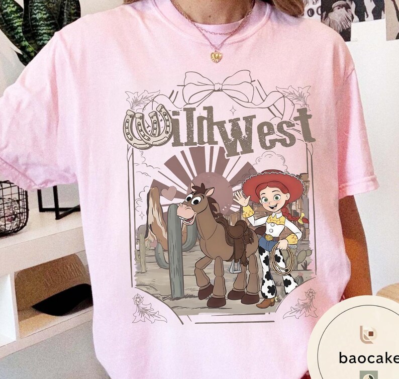 Pu&ograve; includere: T-shirt rosa chiaro con un design a cartoni animati a tema Far West. L'illustrazione include un cavallo, una cowgirl e il testo "Wild West". Il design &egrave; ambientato su uno sfondo desertico con cactus.