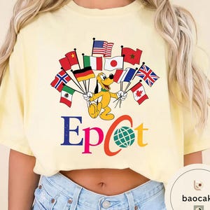 May include: Pale yellow t-shirt with a cartoon Pluto character holding flags from different nations. The word "Epcot" is printed in blue and orange, with a globe graphic. The shirt is a light yellow colour.