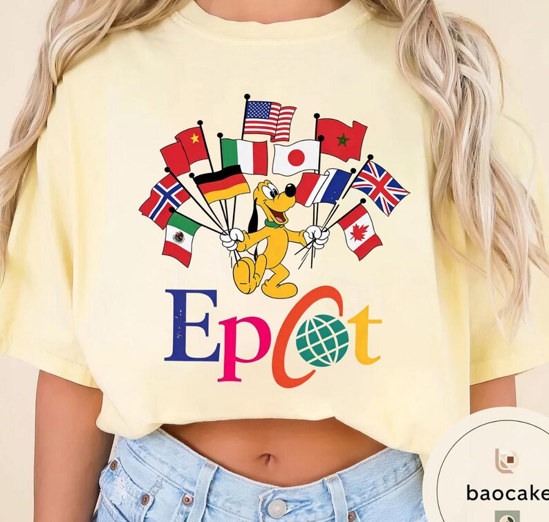 May include: Pale yellow t-shirt with a cartoon Pluto character holding flags from different nations. The word "Epcot" is printed in blue and orange, with a globe graphic. The shirt is a light yellow colour.