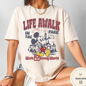 May include: Cream-colored t-shirt featuring a graphic of Mickey Mouse in front of a castle, with the text "LIFE AWALK IN THE PARK" and "Walt Disney World" in red and blue. The shirt has short sleeves.