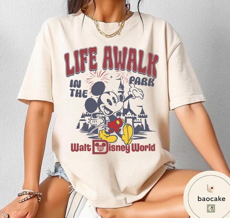 May include: Cream-colored t-shirt featuring a graphic of Mickey Mouse in front of a castle, with the text "LIFE AWALK IN THE PARK" and "Walt Disney World" in red and blue. The shirt has short sleeves.