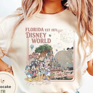 May include: A cream-colored crewneck sweatshirt featuring a graphic print with the text FLORIDA EST 1971 DISNEY WORLD and The Most Magical Place On Earth. The design includes illustrations of Disney characters, a castle, and a geodesic dome.
