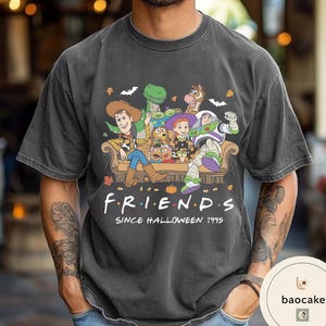 May include: A charcoal grey short-sleeve t-shirt featuring a graphic of Toy Story characters sitting on a couch. The text reads F R I E N D S SINCE HALLOWEEN 1995. The design includes autumn leaves, bats, and spiderwebs surrounding the characters.