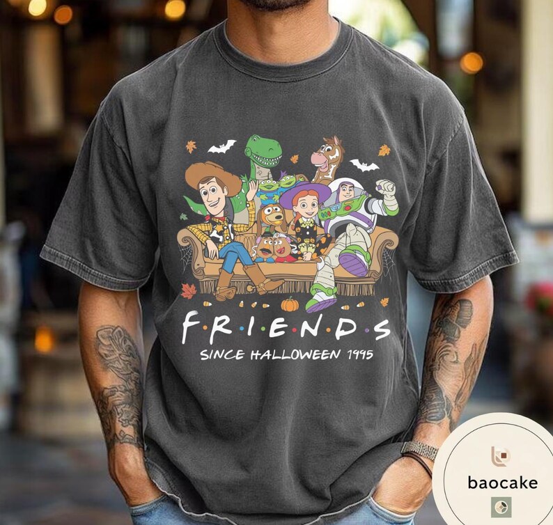 May include: A charcoal grey short-sleeve t-shirt featuring a graphic of Toy Story characters sitting on a couch. The text reads F R I E N D S SINCE HALLOWEEN 1995. The design includes autumn leaves, bats, and spiderwebs surrounding the characters.