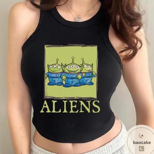 K&ouml;nnte beinhalten: Ein schwarzes &auml;rmelloses Tanktop mit einem Grafikdruck von drei gr&uuml;nen, drei&auml;ugigen Au&szlig;erirdischen in blauen Anz&uuml;gen innerhalb eines hellgr&uuml;nen Quadrats. Das Wort ALIENS ist in gro&szlig;en, hellgr&uuml;nen Gro&szlig;buchstaben unter der Grafik gedruckt.
