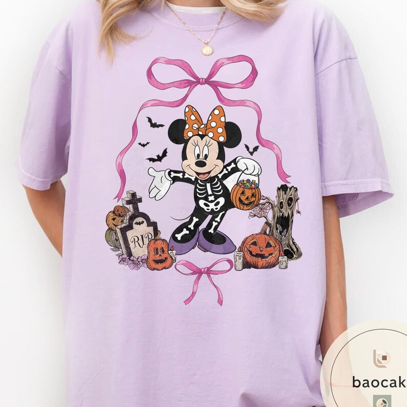 May include: A light purple t-shirt featuring a Halloween-themed Minnie Mouse design. Minnie is dressed as a skeleton, holding a jack-o'-lantern. The design includes pumpkins, bats, a tombstone, and bows. The shirt is a comfortable, oversized fit.