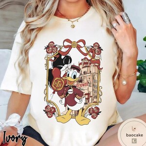 May include: Ivory-colored t-shirt featuring a cartoon of Donald Duck in a maroon bellhop uniform, holding a balloon. The graphic includes the Hollywood Tower Hotel and a decorative frame with roses and a bow.