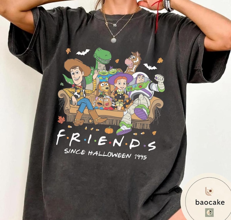May include: A charcoal grey t-shirt featuring a graphic print of Toy Story characters sitting on a sofa. The text reads F R I E N D S SINCE HALLOWEEN 1995. The design includes bats, pumpkins, and autumn leaves, with Woody, Buzz Lightyear, and others.