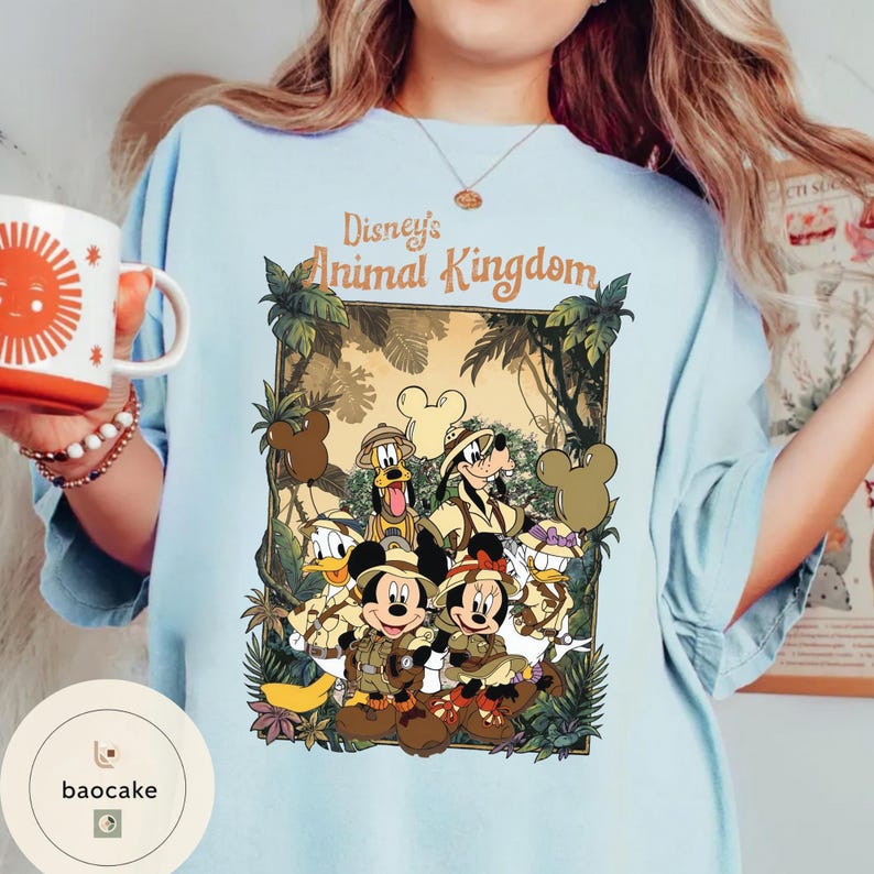 May include: Light blue t-shirt featuring a colorful graphic of Disney's Animal Kingdom characters, including Mickey and Minnie Mouse, Donald Duck, Goofy, and Pluto. The text "Disney's Animal Kingdom" is displayed above the characters. The shirt has short sleeves.