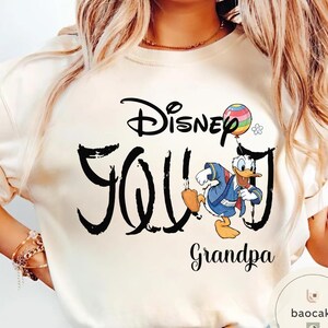 Puede incluir: Sudadera color crema con la inscripci&oacute;n "Disney Grandpa" en escritura negra, con una imagen de dibujos animados de Pato Donald sosteniendo un globo colorido. La palabra "Disney" est&aacute; arqueada sobre la palabra "Grandpa".