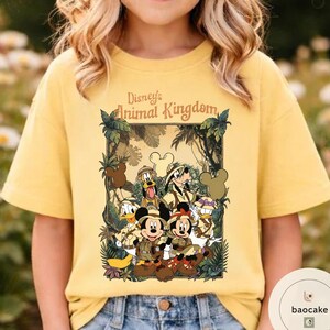 May include: Yellow t-shirt featuring a graphic of Disney's Animal Kingdom characters, including Mickey and Minnie Mouse, Donald Duck, and Goofy. The design is framed with jungle foliage and the text "Disney's Animal Kingdom".