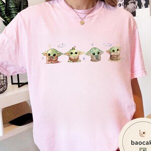 May include: A light pink t-shirt featuring four cartoon illustrations of a green alien-like character. Each character is in a different pose, with a variety of brown and tan clothing. The design includes small star and cross accents.