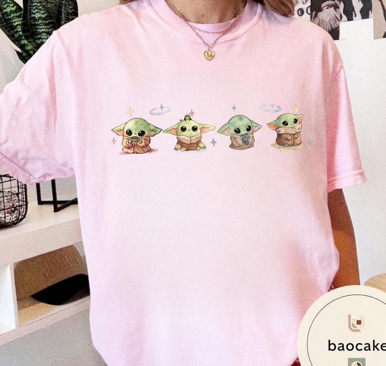 May include: A light pink t-shirt featuring four cartoon illustrations of a green alien-like character. Each character is in a different pose, with a variety of brown and tan clothing. The design includes small star and cross accents.