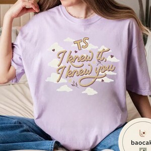 K&ouml;nnte beinhalten: Ein hellviolettes T-Shirt mit einem Grafikdruck aus wei&szlig;en Wolken und dem Text "TS I knew it, I knew you" in einer beigefarbenen Schrift im Seil-Stil. Das Design enth&auml;lt kleine dunkelbraune Herzen und dekorative Zweige zwischen den Wolken.