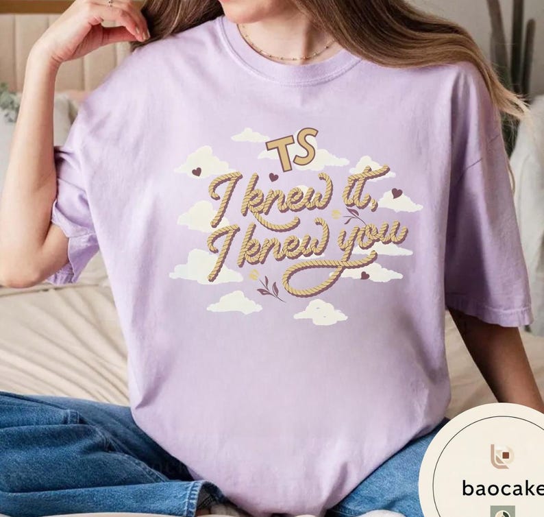 K&ouml;nnte beinhalten: Ein hellviolettes T-Shirt mit einem Grafikdruck aus wei&szlig;en Wolken und dem Text "TS I knew it, I knew you" in einer beigefarbenen Schrift im Seil-Stil. Das Design enth&auml;lt kleine dunkelbraune Herzen und dekorative Zweige zwischen den Wolken.