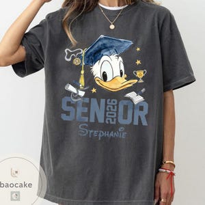 May include: Dark gray t-shirt featuring Donald Duck wearing a graduation cap. The shirt has the text "SENIOR 2026" and a personalized name. Additional graphics include a diploma, trophy, and stars.