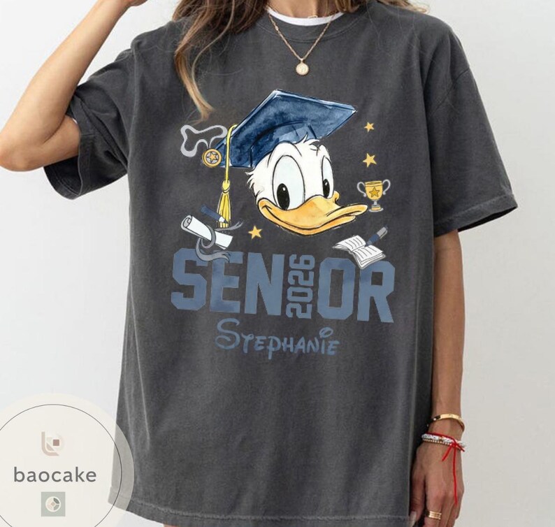 May include: Dark gray t-shirt featuring Donald Duck wearing a graduation cap. The shirt has the text "SENIOR 2026" and a personalized name. Additional graphics include a diploma, trophy, and stars.