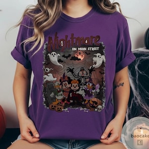May include: Purple t-shirt featuring a Halloween-themed graphic with the text "Nightmare on Main Street." The design includes cartoon characters, ghosts, pumpkins, and a full moon, creating a spooky atmosphere.