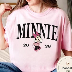 May include: A light pink t-shirt featuring the word "MINNIE" in black block letters. Below the word is a graphic of Minnie Mouse in a pink dress and bow. The numbers "20" and "26" are on either side of the graphic.