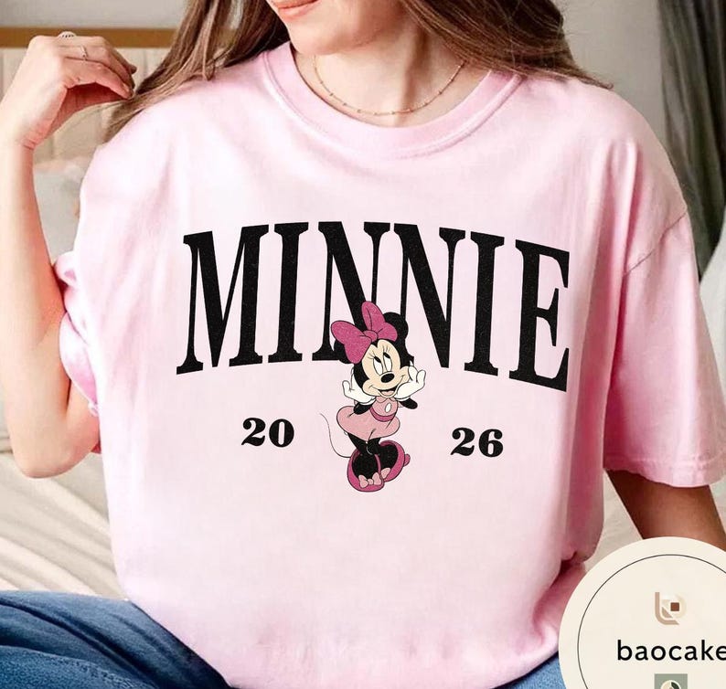 May include: A light pink t-shirt featuring the word "MINNIE" in black block letters. Below the word is a graphic of Minnie Mouse in a pink dress and bow. The numbers "20" and "26" are on either side of the graphic.