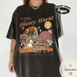 Op de afbeelding: Donkergrijs T-shirt met een Halloween-thema Disney World-afbeelding. Het ontwerp bevat pompoenen, geesten en een stripfiguur voor een kasteel. De tekst op het shirt luidt "Florida Disney World" en "The Most Scariest Place On Earth."