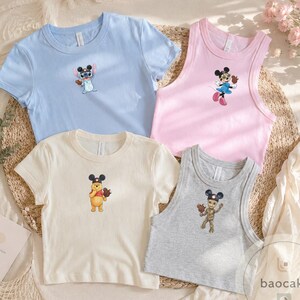 May include: A collection of four shirts featuring Disney character graphics. The set includes a light blue t-shirt with Stitch, a pink tank top with Minnie Mouse, a cream t-shirt with Winnie the Pooh, and a gray tank top with Groot, all wearing mouse ears.