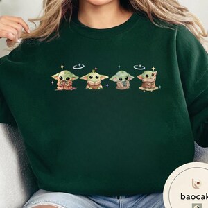 May include: Dark green sweatshirt featuring a row of four Grogu illustrations. Each Grogu is depicted in a different pose, with a halo and stars. The sweatshirt is a solid color, with the baocake logo in the bottom right corner.