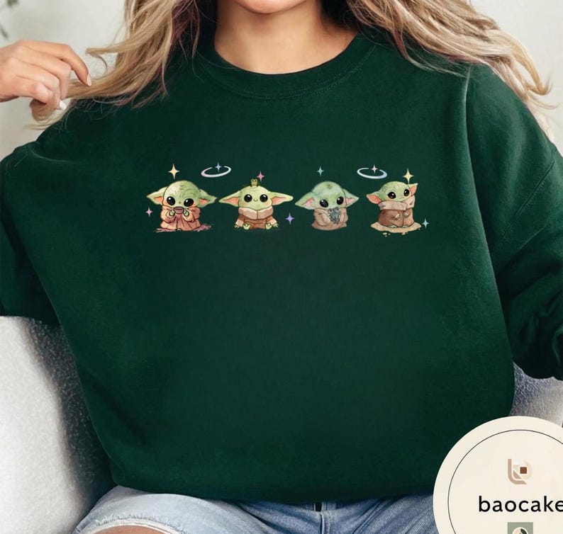 May include: Dark green sweatshirt featuring a row of four Grogu illustrations. Each Grogu is depicted in a different pose, with a halo and stars. The sweatshirt is a solid color, with the baocake logo in the bottom right corner.