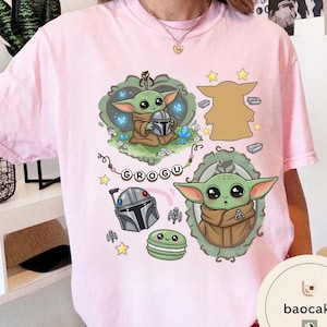 May include: A light pink t-shirt featuring a graphic print of a green alien character, a silver helmet, a green macaron, and stars. The design includes a string of beads spelling the name GROGU, along with two framed illustrations of the character.