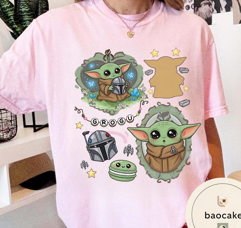 May include: A light pink t-shirt featuring a graphic print of a green alien character, a silver helmet, a green macaron, and stars. The design includes a string of beads spelling the name GROGU, along with two framed illustrations of the character.