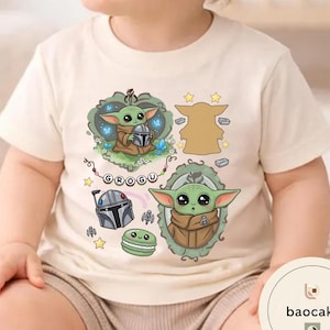 May include: A cream-colored short-sleeve t-shirt featuring a graphic print of the character Grogu. The design includes two illustrations of the character, a silver helmet, a green macaron, stars, and a banner with the text GROGU in black letters.
