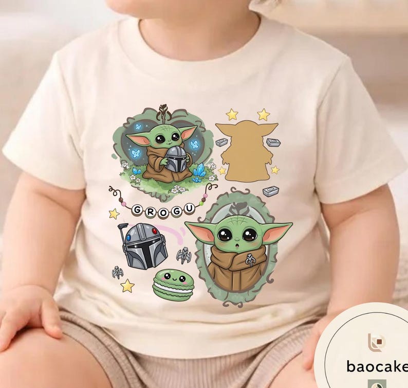 May include: A cream-colored short-sleeve t-shirt featuring a graphic print of the character Grogu. The design includes two illustrations of the character, a silver helmet, a green macaron, stars, and a banner with the text GROGU in black letters.