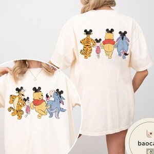 May include: Cream-colored t-shirt featuring a cartoon illustration of Winnie the Pooh, Tigger, Piglet, and Eeyore. The front and back of the shirt display the characters in a line, wearing mouse ears. The design is printed on a soft, oversized tee.