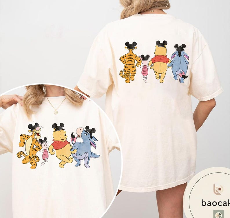 May include: Cream-colored t-shirt featuring a cartoon illustration of Winnie the Pooh, Tigger, Piglet, and Eeyore. The front and back of the shirt display the characters in a line, wearing mouse ears. The design is printed on a soft, oversized tee.