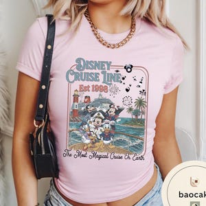 Puede incluir: Una camiseta rosa claro con un dise&ntilde;o de Disney Cruise Line. El dise&ntilde;o incluye a Mickey Mouse, Minnie Mouse, Donald Duck y otros personajes en un crucero, con el texto "Disney Cruise Line Est 1998" y "The Most Magical Cruise On Earth."