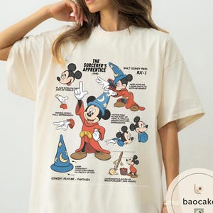 May include: Cream-colored t-shirt featuring a graphic of Mickey Mouse from "The Sorcerer's Apprentice" with text and illustrations. The design includes Mickey in a sorcerer's outfit, a blue hat, and various poses. The text includes "The Sorcerer's Apprentice -1940- Walt Disney Prod RX-1".