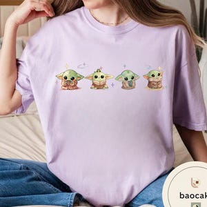 May include: A light purple t-shirt featuring four cartoon images of a green alien character. Each character is in a different pose, with a variety of outfits and accessories. The shirt is made of a soft material.