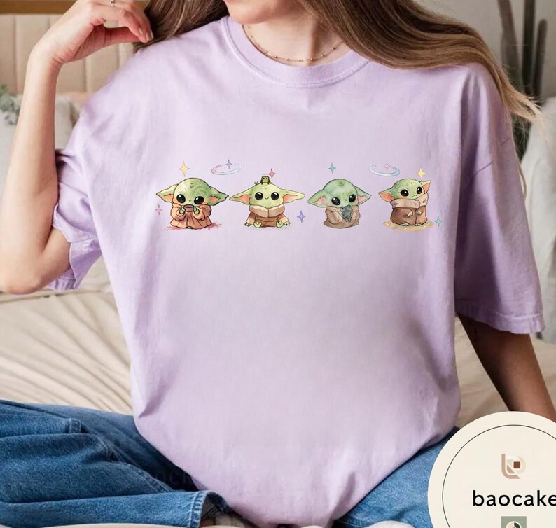 May include: A light purple t-shirt featuring four cartoon images of a green alien character. Each character is in a different pose, with a variety of outfits and accessories. The shirt is made of a soft material.