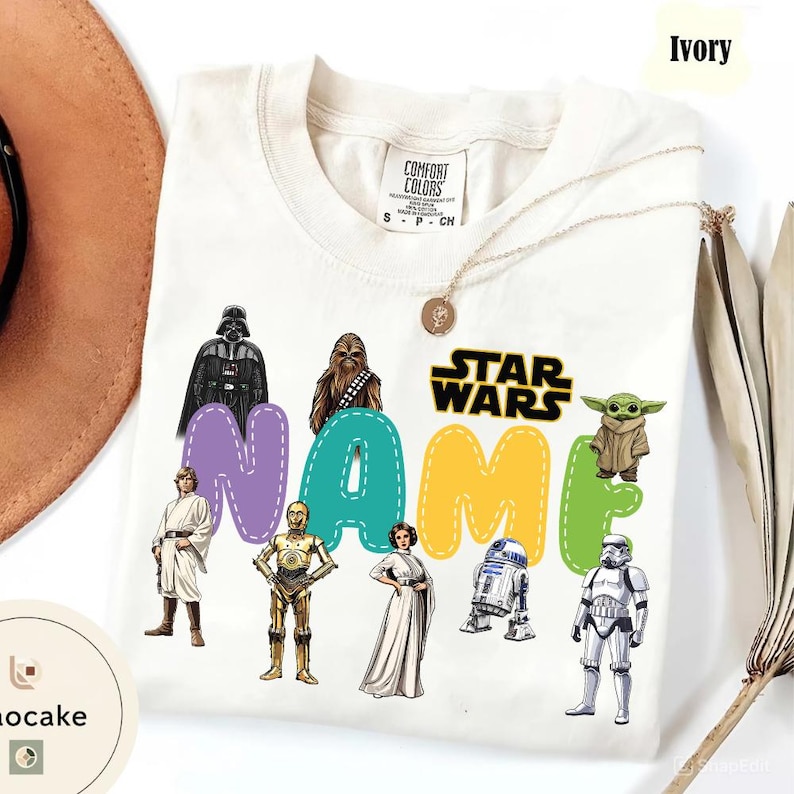 May include: Ivory-colored t-shirt featuring Star Wars characters and the text "NAME" in colorful letters. The shirt includes images of Darth Vader, Chewbacca, Yoda, and other characters from the Star Wars franchise. A gold necklace is draped on the shirt.