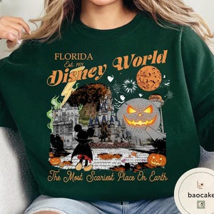May include: Dark green sweatshirt with a Halloween-themed graphic. The design includes text that reads "FLORIDA Disney World Est. 1971" and "The Most Scariest Place On Earth". The graphic features a Mickey Mouse character, a pumpkin, and a castle.