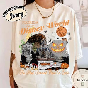 May include: Ivory-colored t-shirt featuring a Halloween-themed Disney World graphic. The design includes a pumpkin, Mickey Mouse in a cape, and the text "The Most Scariest Place On Earth." The shirt also has the words "Florida" and "Disney World."