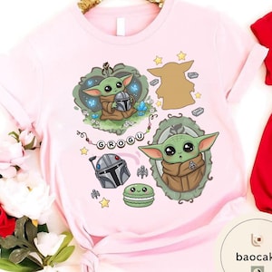 May include: A light pink t-shirt featuring a graphic print of a green alien character in a brown robe, a silver helmet, a green macaron, and a string of beads spelling GROGU. The design includes various small stars, blue crystals, and a silhouette.