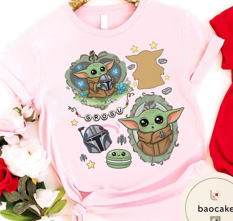 May include: A light pink t-shirt featuring a graphic print of a green alien character in a brown robe, a silver helmet, a green macaron, and a string of beads spelling GROGU. The design includes various small stars, blue crystals, and a silhouette.